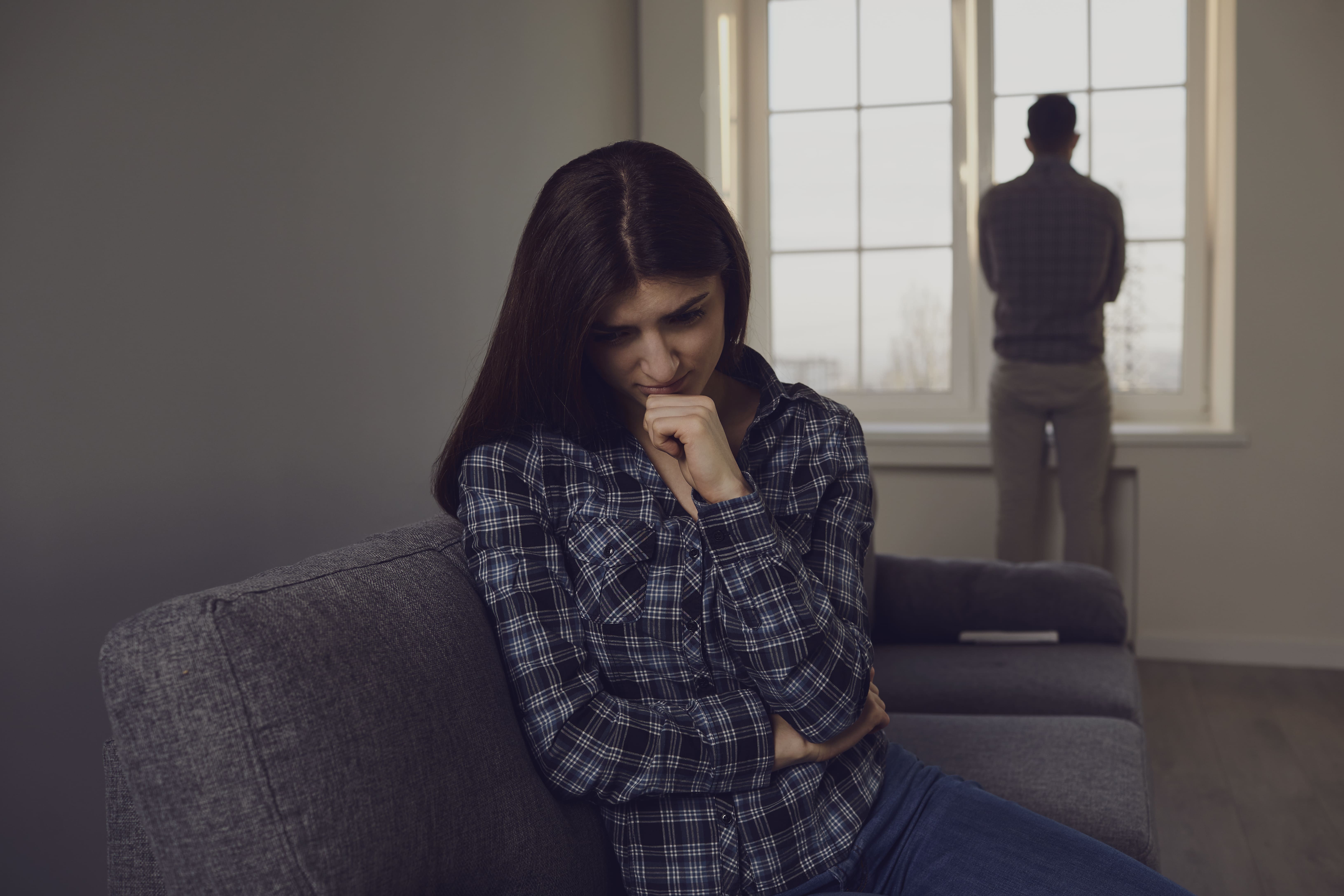 Couple en conflit, femme assise pensive, homme regardant par la fenêtre.
